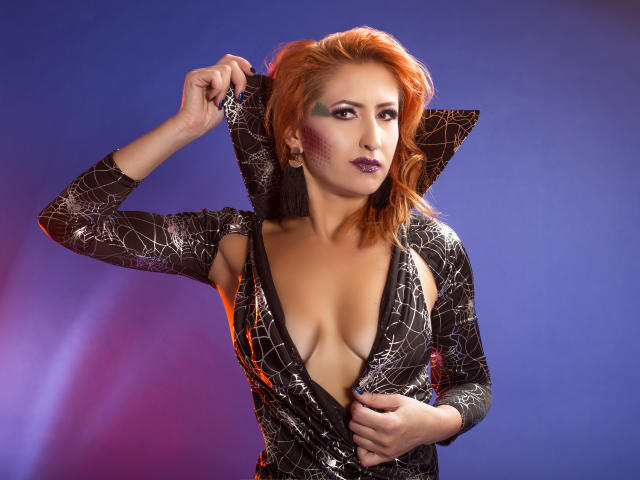 IvyFox con i capelli rossi, scollatura profonda in un abito nero con motivo a ragnatela, che tiene sollevato il colletto, trucco agli occhi viola, rossetto viola, sfondo blu, mano sul petto, che guarda verso l'alto..