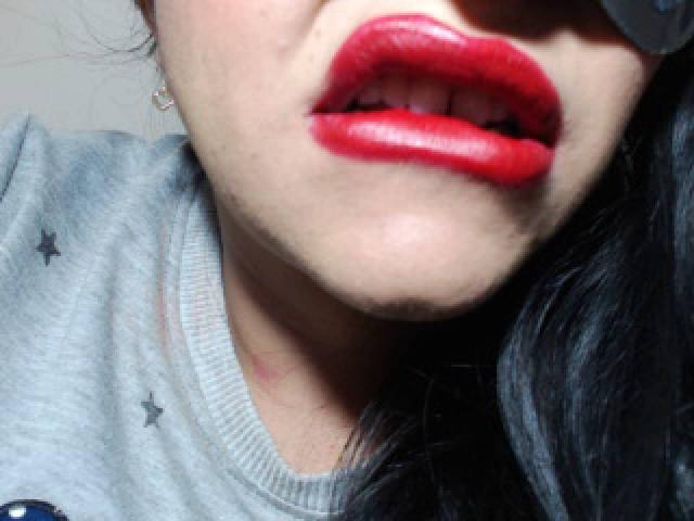 LuluFrancaise's glossy red lips are slightly open, showing her white teeth. She's wearing a gray shirt with small star patterns, and her black hair is partially visible.