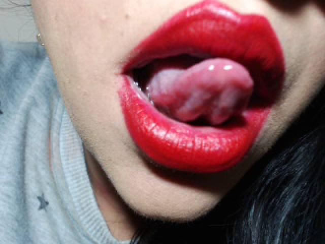 LuluFrancaise's glossy red lips are open wide, showing her tongue coated in white fluid. Her tongue is slightly curled, with visible saliva. She's wearing a grey shirt.