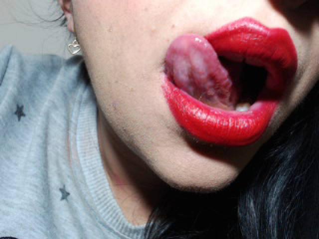LuluFrancaise's face with glossy red lipstick, tongue out, slightly open mouth, showing a bit of her teeth, wearing a gray shirt, and small hoop earrings.