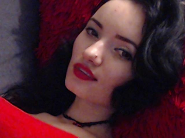 MissVanesa tiene lápiz labial rojo, cabello oscuro y ondulado, y un collar negro. Está acostada sobre una superficie roja y esponjosa, con una expresión sensual, ligeramente de boca abierta..