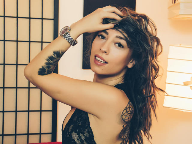 PoxyVibe with wavy brown hair, black lace top, and feather tattoo on arm, looking back with hand in hair, smiling, wearing a silver watch. Warm indoor lighting.