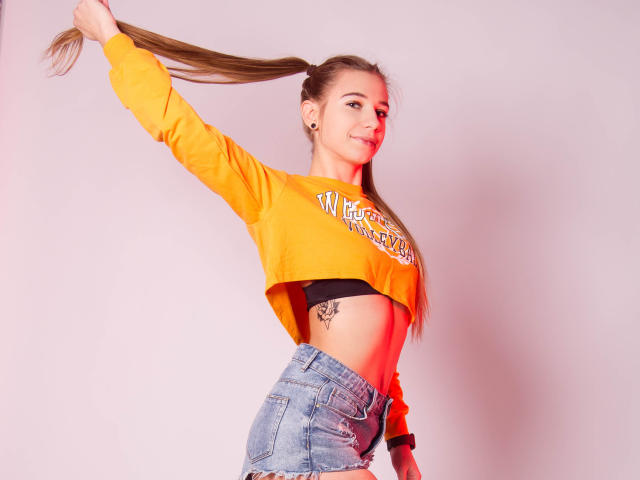 LucyBestCandy with long brown hair, yellow crop top, black bra, tattoo under right boob, blue jeans, holding hair, smiling, light pink background, sexy, slim, cute.