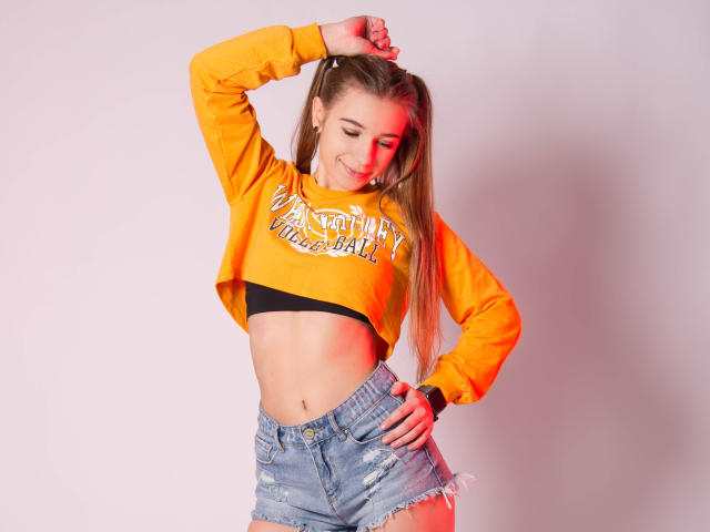 LucyBestCandy, con lunghi capelli biondi, indossa un top corto arancione, un reggiseno nero e pantaloncini di jeans strappati, sorridendo, con una mano tra i capelli, mostrando il suo addome tonico e il suo sedere carino..