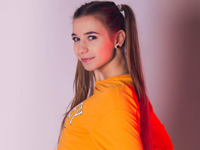 LucyBestCandy, con lunghe trecce marroni, piccoli orecchini neri e un sorriso carino, indossa una maglietta arancione e ha un piccolo punto rosso sulla guancia..