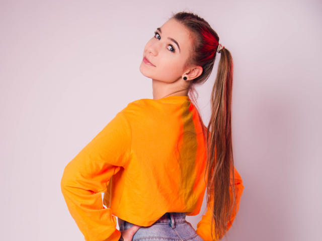 LucyBestCandy, avec une longue queue de cheval brune, portant une chemise orange à manches longues, un jean bleu et de petites boucles d'oreilles, regardant par-dessus son épaule avec un léger sourire..