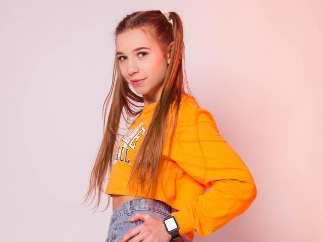 LucyBestCandy, con largas coletas marrones, un top naranja y jeans azules, mostrando su trasero con una mano en la cadera, y usando un reloj inteligente negro..