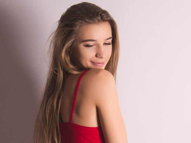 LucyBestCandy mit langen, braunen Haaren, einem roten Spaghetti-Top, lächelt leicht und schaut über ihre Schulter, wobei ihr nackter Rücken und ihre glatte Haut zu sehen sind. Ihre Brüste sind klein und niedlich..