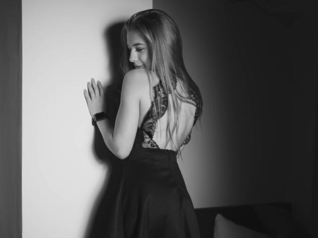 LucyBestCandy, con lunghi capelli biondi, indossa un abito nero con dettagli in pizzo sulla schiena, è in piedi contro un muro, con le mani alzate, guarda in basso, indossa un braccialetto nero, foto in bianco e nero, luce soffusa..
