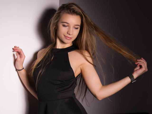 LucyBestCandy with long brown hair, wearing a black halter dress, smiling, holding her hair, light skin, black bracelet, standing against a white and dark gray wall.