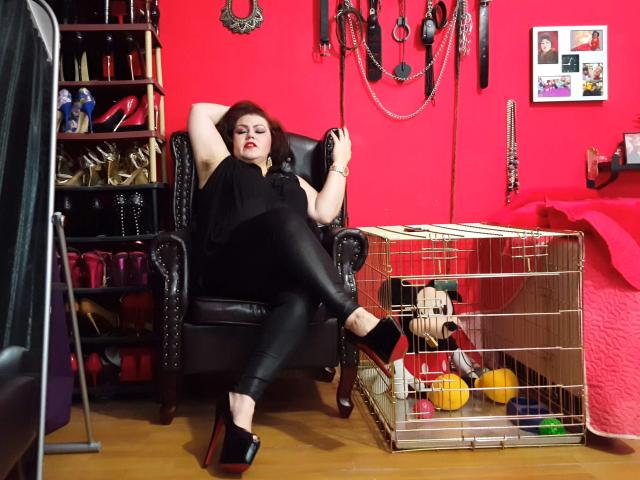 KaryQueen with red lipstick, black shirt, and black leather pants, lounging in a black studded chair, phone in hand, high heels on, cage with toys on wooden floor.