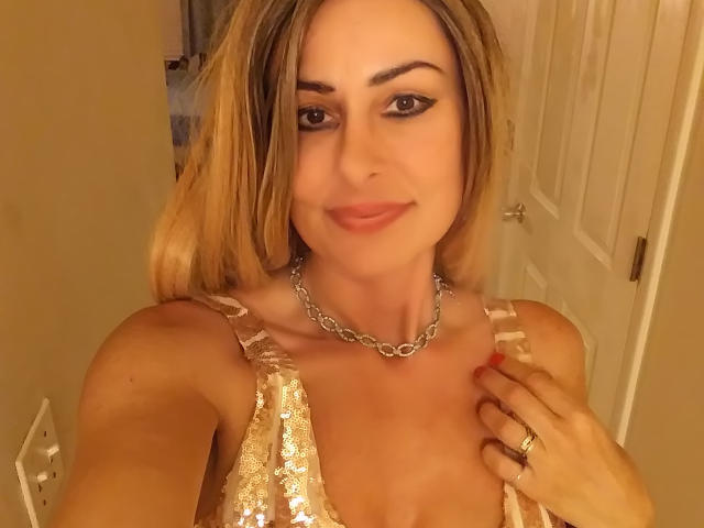 NoraNoirX has blonde hair, wearing a sequined gold top and silver chain necklace, smiling with a hand on her chest, showing a ring. Boobs, small, visible under top.