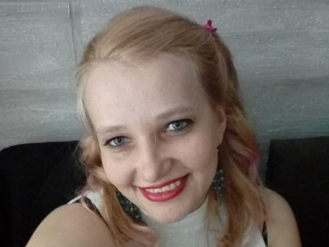 A AnytkaKiss tem cabelo loiro claro, olhos azuis e batom vermelho, sorrindo para a câmera com um prendedor de cabelo rosa no lado direito, vestindo uma blusa branca..