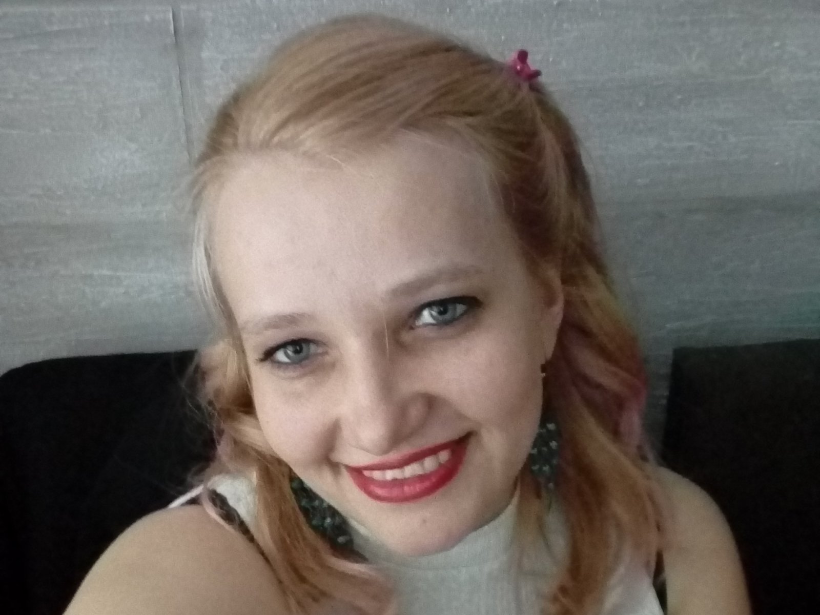 AnytkaKiss tiene el cabello rubio claro, ojos azules y lápiz labial rojo, sonriendo a la cámara con una pinza para el cabello rosa en el lado derecho, y lleva una blusa blanca..