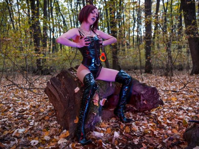ClaraDomme with red bob, wearing black shiny latex bodysuit, orange ring, and thigh-high boots, sitting on tree stump, hands on tits, forest floor, autumn leaves, seductive pose.