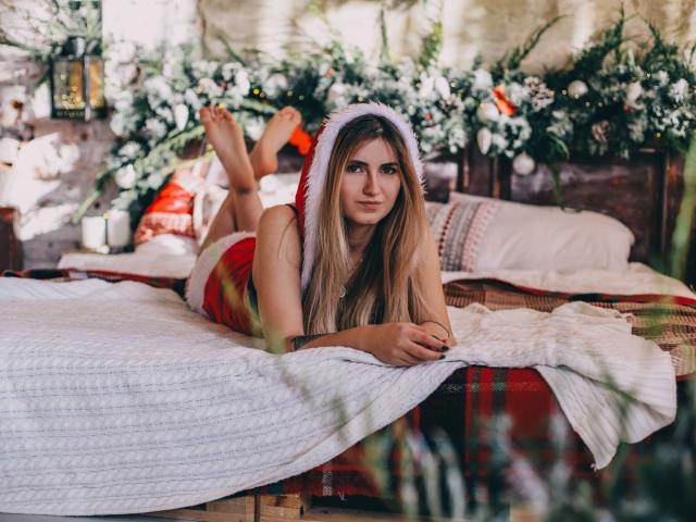ShyNaomi met lang blond haar, een kerstmuts op, liggend op een bed, benen omhoog, rode jurk, wit laken, geruit deken, kerstdecoratie, blote voeten, schattig, sexy..