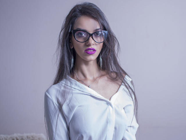 KatherineBisou with long dark hair, glasses, purple lipstick, white button-up shirt, and a subtle necklace, looking serious. Her tits are slightly visible under the shirt.
