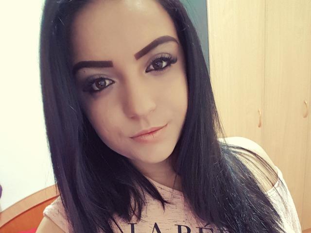 Adelinee ha i capelli neri e lisci, le sopracciglia marroni, e guarda la fotocamera con un leggero sorriso. Indossa una camicia rosa, con il naso visibile e un trucco leggero..