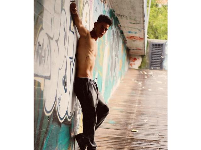 Shirtless DerrickBigX with a toned, muscular upper body leans against a graffiti-covered wall, wearing loose black pants, standing on a wet wooden bridge, looking down.