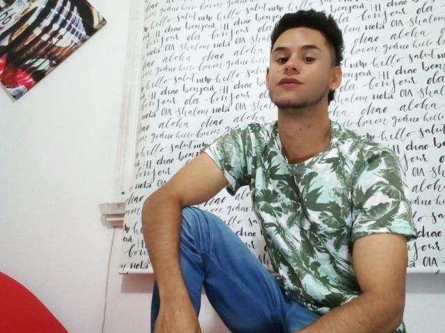 DerrickBigX, vestindo uma camisa verde com estampa de folhas de palmeira e calças azuis, está sentado com uma expressão relaxada, ligeiramente irritada. Seus cabelos curtos e escuros e sua pele clara são visíveis..