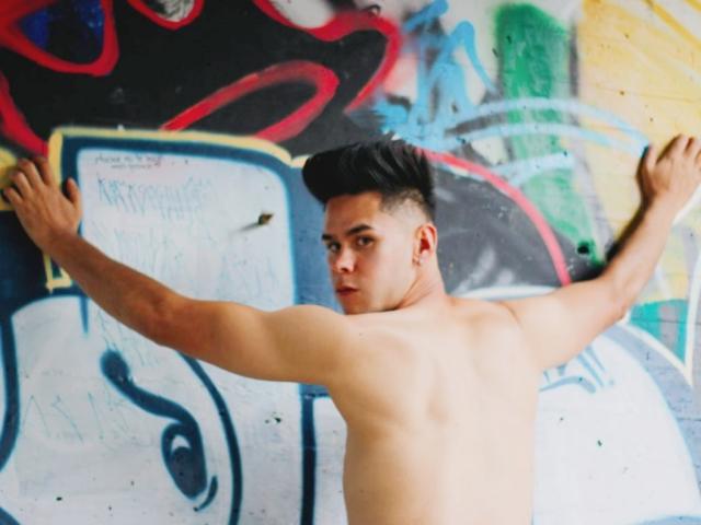 DerrickBigX, sem camisa, musculoso, cabelo preto, olhando para trás, braços abertos, em frente a grafites coloridos, expressão séria, tom de pele claro, costas nuas, levemente suado, cabelo curto, ambiente urbano..