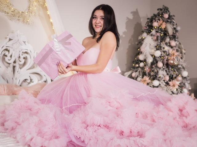 CuttieEyesX con grandi seni, in un abito rosa, soffice e senza spalline, che tiene regali rosa, seduta su un letto, sorridendo, accanto a un albero di Natale decorato..