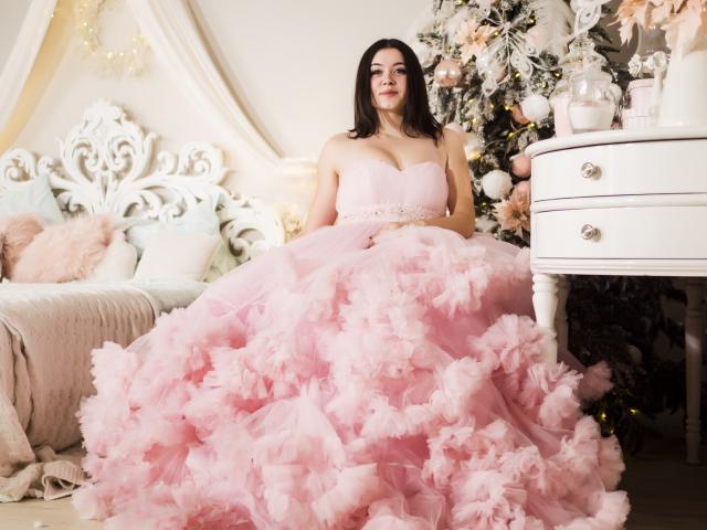 CuttieEyesX with pink feathered ballgown, showing off her big boobs and fluffy ass, standing in front of a white dresser with a Christmas tree to the right.