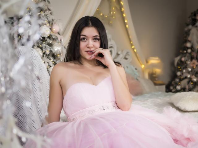 CuttieEyesX with long dark hair, wearing a pink strapless dress with a fluffy skirt, sits on a bed, touching her lips, showing her tits and a cute smile.