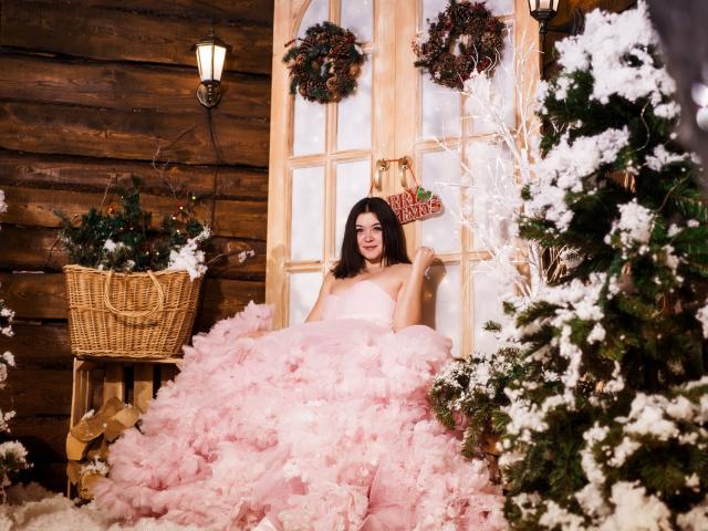 CuttieEyesX trägt ein voluminöses, pinkes Kleid, die Brüste sind kaum sichtbar. Sie sitzt zwischen zwei mit Schnee bedeckten Weihnachtsbäumen, im Hintergrund sind eine Holzwand, ein Korbkorb, eine Laterne und zwei Kränze zu sehen..