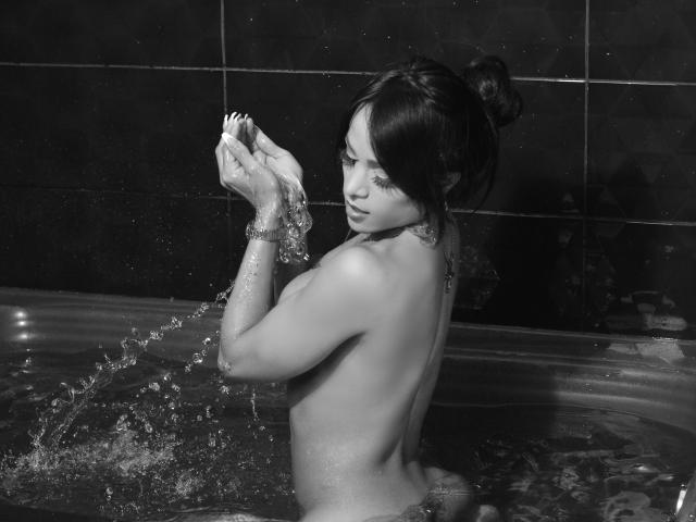 LorentsTS is in a black-and-white photo, topless in a bathtub, holding water in her hands, with her tits and dark hair in a bun, looking sideways.