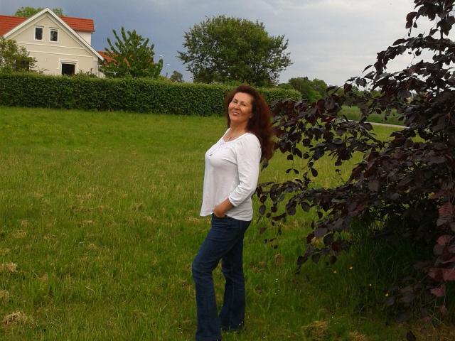 JuicyXSandra met golvend bruin haar, een wit shirt en blauwe jeans staat in een grasveld, met haar handen in haar zakken, lachend, naast een boom met donkere bladeren, onder bewolkte luchten..