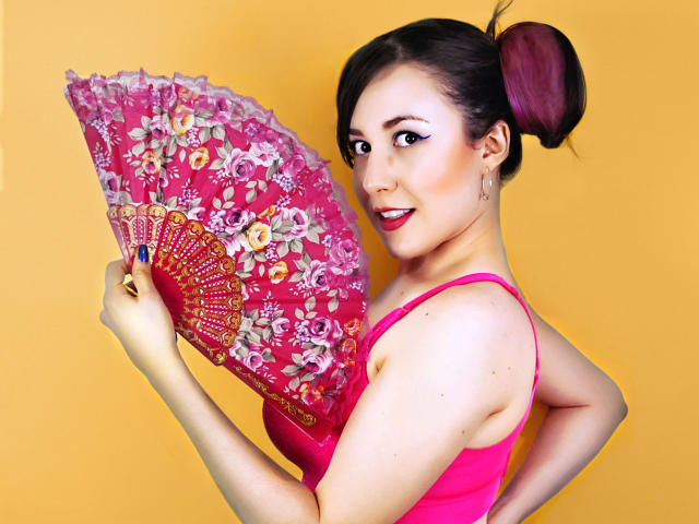MabyLegrand with pink floral fan, holding it in front of her tits, wearing a pink dress, yellow background, dark brown hair in updo, smiling, red lipstick, earrings.