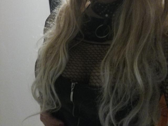 RoxannaTS mit langen, welligen blonden Haaren, die ein schwarzes Netzoberteil trägt, durch das ihre Brüste sichtbar sind, und eine schwarze Jacke mit Reißverschluss, in einer schwach beleuchteten Umgebung..