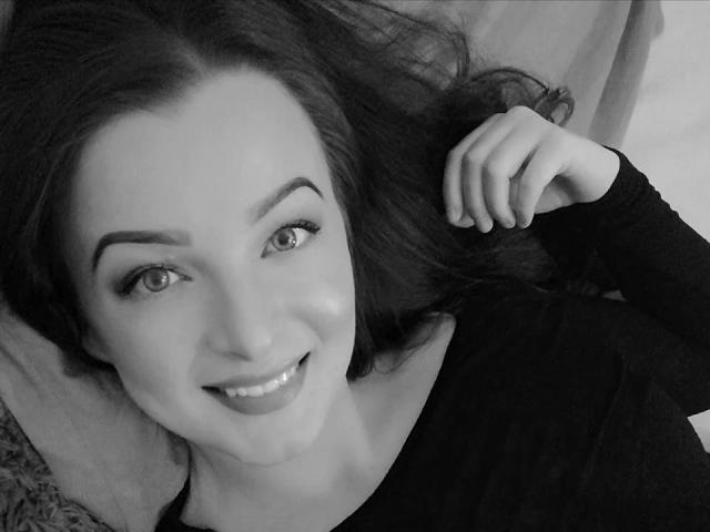 Autoportrait en noir et blanc de StellaLee, avec des cheveux foncés, souriante, portant un haut noir à manches longues, allongée, la main droite touchant ses cheveux. Les seins ne sont pas visibles, le focus est sur le visage..