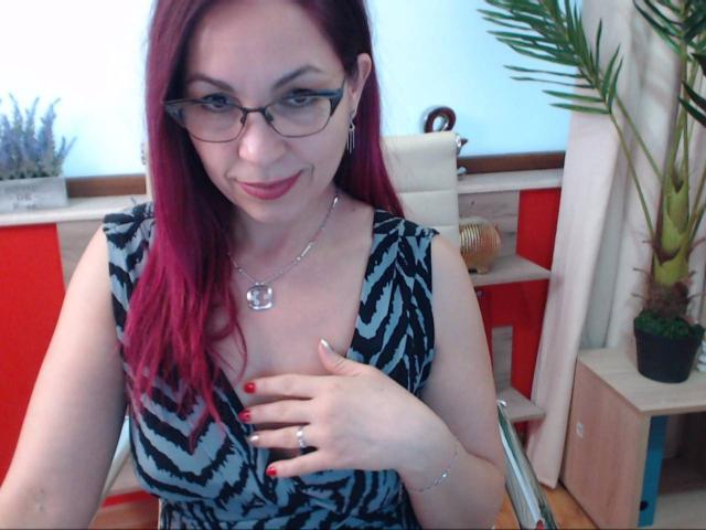 DirttyMature with red hair, glasses, and a black-and-blue patterned dress, covering her tits with her hand, red nails, smiling, necklace, and a ring, in a chair.