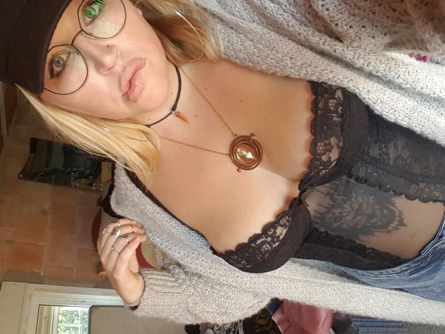 Shushaina mit grünen Augen, Brille, schwarzer Spitzen-BH, schwarzem Spitzen-Korsett, grauem Cardigan, Mütze, Halskette und Jeans, die ihre Brüste und ihren tätowierten Bauch zeigt..