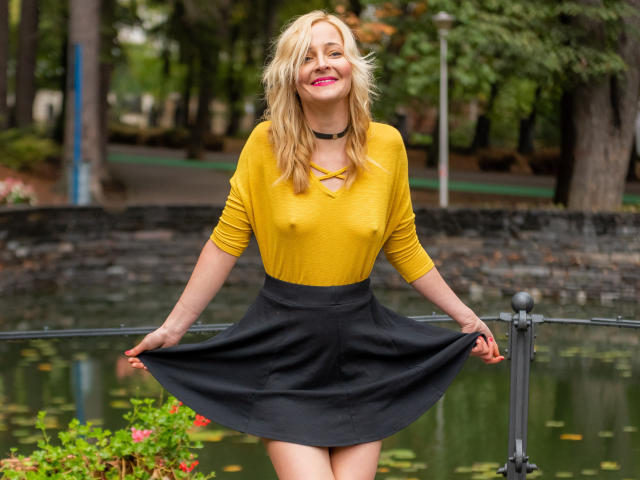 SoleildeNuit has blonde hair, pink lipstick, yellow top, showing nipples, black skirt, holding skirt, black choker, smiling, standing by a pond, hands on skirt, green background.