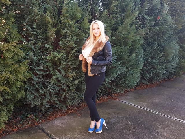 Dajla69 met lang blond haar, een zwarte leren jas, een bruine bontvest, zwarte broek en blauwe hoge hakken, staat voor hoge groene bomen en glimlacht naar de camera..