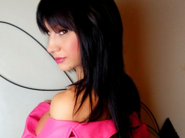Cassandra Michelli mit langen, schwarzen Haaren, pinkem Lippenstift und einem pinken Top mit einem Schulterauschnitt, die über ihre Schulter schaut und dabei ihre nackten Schultern und einen Hauch ihrer Brüste zeigt..