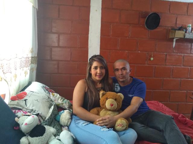 KratussyAtenea mit langen braunen Haaren, die ein schwarzes Oberteil und blaue Jeans trägt, sitzt auf einem Bett mit einem Teddybären, neben einem kahlköpfigen Mann in einem blauen Hemd..