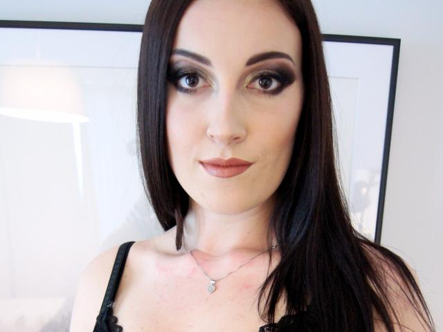 AmandaChilli with long brown hair, dark smoky eye makeup, and pink lipstick, wearing a black lace top and a silver necklace, looking at the camera with a slight smile.