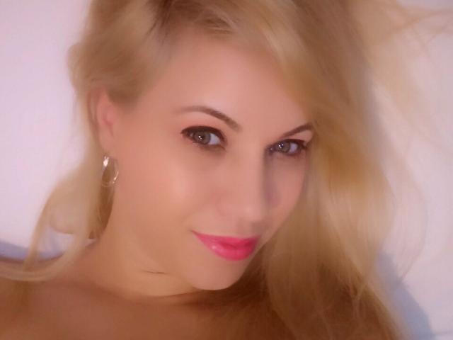 MyaHott has blonde hair, pink lipstick, and is smiling with hoop earrings. Her makeup is subtle, and she has a cute, sexy look. Her tits are not visible.