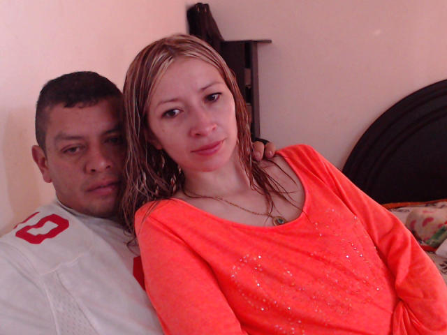 Casal latino sensual com cabelo loiro platinado, seios pequenos em uma camisa laranja neon, sentada ao lado de um homem latino com cabelo curto e escuro, vestindo uma camisa branca, em uma cama..