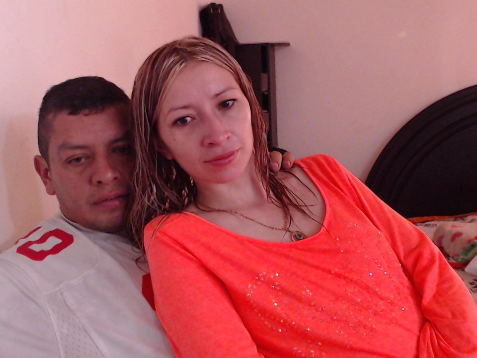 Un couple attrayant, une femme latina aux cheveux blonds et clairs, avec de petites seins, portant une chemise orange néon, est assise à côté d'un homme latino aux cheveux courts et foncés, vêtu d'une chemise blanche, sur un lit..