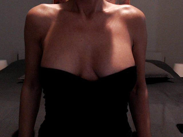 PrettyEllen's bare shoulders and black strapless top show her small, perky tits and cleavage, with her skin glowing in dim light. She stands in a dark bedroom.