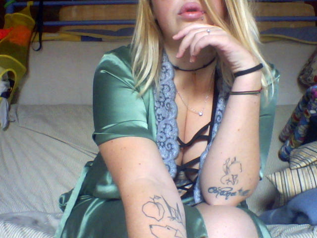 Shushaina mit blonden Haaren, einem grünen Seidenkleid, Spitzenbesatz, einem schwarzen Trägeroberteil, Tattoos an den Armen, einem Lierepiercing, einer Hand am Kinn, sitzend auf dem Bett, einer Choker-Kette und einem Ring am Finger..