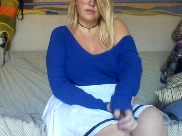 Shushaina, avec ses cheveux blonds, ses lunettes, son pull bleu à une épaule, sa jupe blanche, son collier, tenant une télécommande, assise sur le lit, les jambes jointes, avec une partie de ses parties intimes légèrement visible, en tenue décontractée, détendue..