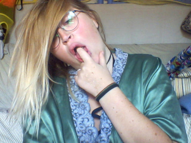 Shushaina with blonde hair, glasses, and green satin shirt, sucks her finger, her mouth slightly open, showing a bit of her tongue and teeth. Her hand's on her chin.