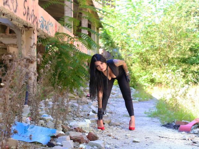 VanessaOdette, penchée en avant, vêtue d'un pantalon noir et de talons rouges, avec de longs cheveux noirs et un tatouage sur le bras, regardant vers le bas, debout sur un sentier rocheux avec de la verdure et un bâtiment abandonné..