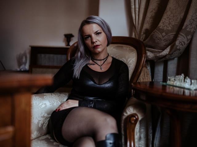 VanessaOdette, avec ses cheveux gris, son haut en cuir noir, sa jupe noire et ses bottes noires, assise sur une chaise ornée, les jambes croisées, avec une expression sérieuse, un collier noir, et un tatouage visible sur la poitrine..